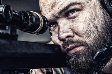 Army sniper with bearded, dirty face aiming with optical telescopic sight on sniper rifle,...