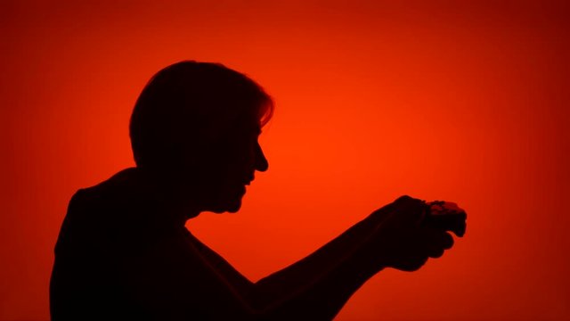 Silhouette of senior woman gamer playing video game online. Female's face in profile with game console on red background. Black contour shadow of grandmother's half-face winning computer game