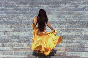 A girl in a gold traditional oriental costume for belly dancing will climb the stairs with her back to the viewer. The wind develops her hair and hem dresses.