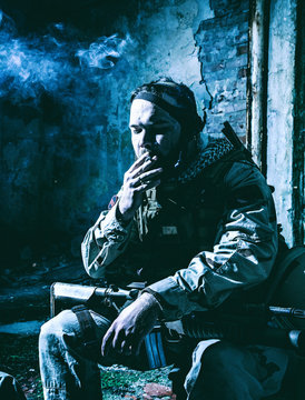 Tired after hard battle army soldier, exhausted with fight Navy Seal rifleman sitting with assault rifle on knees, resting and smoking cigarette in abandoned building, low key, high contrast shoot