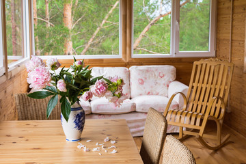 A cozy terrace with furniture - a wooden rocking chair, a sofa, peonies in a vase on the table and wicker chairs with a view of the forest.