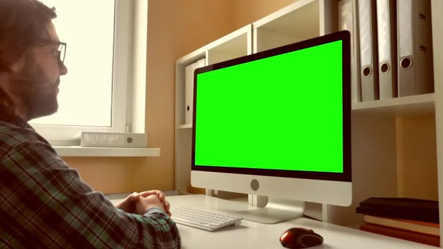 man sitting at workplace paying game on computer in small office. businessman with beard looking on display with green screen. employee in startup company typing on keyboard use pc mouse
