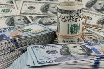 A roll of one hundred dollar bills tied with a rope against the backdrop of scattered dollars.