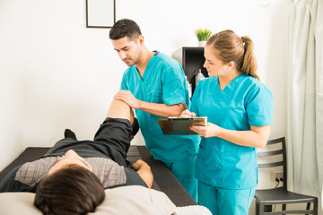 Physiotherapist Examining Customer's Leg While Colleague Noting In Clipboard