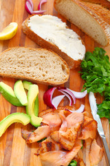 Bread, smoked salmon, avocado, cream cheese, onions and lettuce on a cutting board vertical