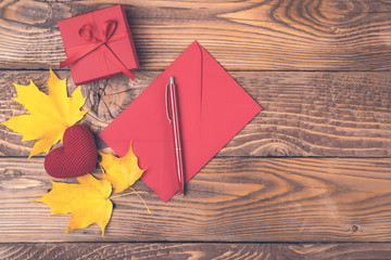 Autumn background with maple leaves, red envelope, pen, gift box, knitted heart on wooden boards. Copy space, top view. Autumn holiday concept. Love in autumn time. Autumn mood. Confession in feelings