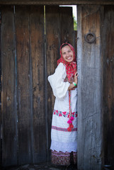 A woman in a Russian folk dress and a scarf standing at the wooden gate and smiling