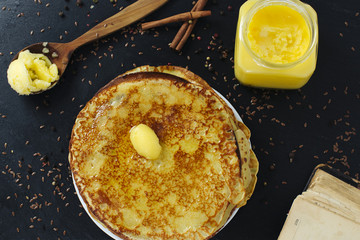 Pancakes are abundantly covered with creamy melted butter on a stone tray and a jar with butter, wooden spoon with butter in it and old book