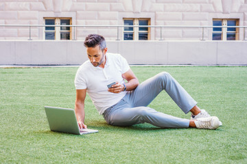 European Man studying, working in New York, with beard, little gray hair, wearing white Polo shirt, gray pants, white sneakers, sitting on green lawn, working on laptop computer, texting on cell phone - Powered by Adobe