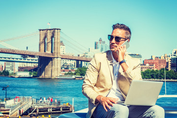 Young European Businessman traveling, working in New York, with beard, little gray hair, wearing beige blazer, sunglasses, sitting by East River, working on laptop computer, talking on cell phone. .