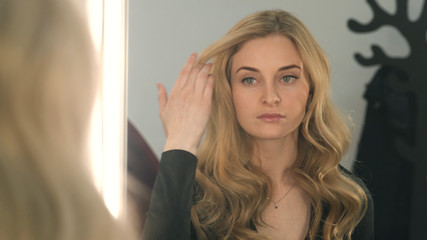 Beautiful blonde girl looks at herself in the mirror and straightens her long hair. Woman corrects her hair before a mirror.