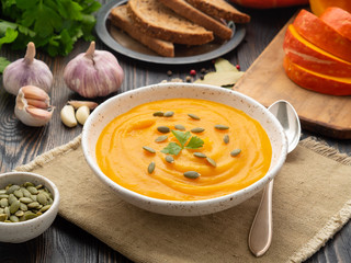pumpkin soup mashed in a white plate, pumpkin seeds, bread, garlic on a dark wooden background,...