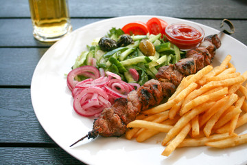  Shashlik or shashlyk of pork, dish of skewered and grilled cubes of meat, with red onion, vegetables, tomatoes, olives and french fries on a wooden table with beer in the background
