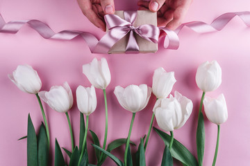 Male hands holding a gift with ribbons over pink background with white tulips. Greeting card or wedding invitation