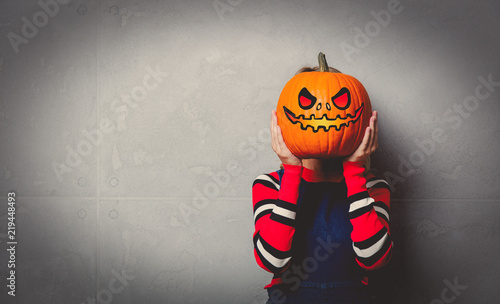 Portrait of a young style blonde girl in red striped sweater with halloween pumpkin on grey background.