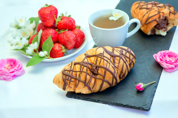 Herbal tea, beautiful pink rose flowers, ripe strawberry and chocolate croissant on the table. Sweet snack. 