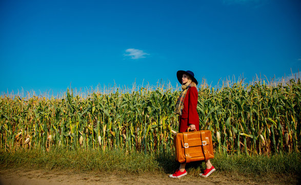 Young girl in coat with suitcase walking along a rural road near cornfield.