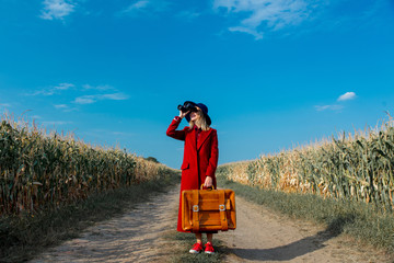 Young girl in coat with suitcase and binoculars on a rural road near cornfield.