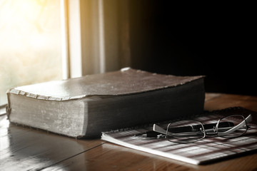 Holy bible with eye glasses and notebook, pen on wooden table with window light in the morning.