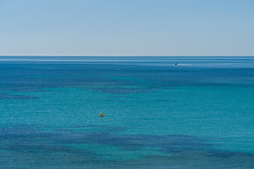 Different blue shades at a beach of the Mediterranean Sea with a yacht at the background on a summer day with clear sky