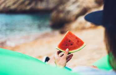 Fototapeta na wymiar girl hipster relax on beach coast and holds in hand a slice of red fresh fruit watermelon on blue sea background, woman on seaside nature eating sweet healthy food, vacation in summer concept