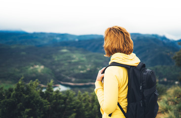tourist traveler with yellow backpack standing on green top on mountain, hiker view from back looking on hills and mountain river lake, girl enjoying panoramic landscape in trip, relax holiday concept