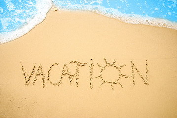 Word VACATION written on the beach sand, with a blue wave