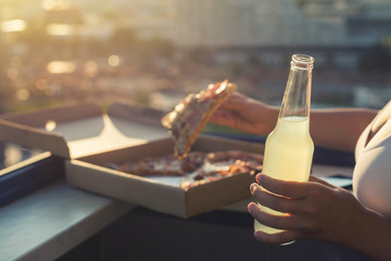 A female in a sports suit holds a huge piece of pizza on sunset and city background.