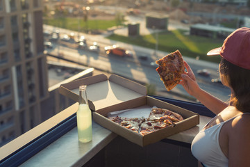 A female in a sports suit holds a huge piece of pizza on sunset and city background.
