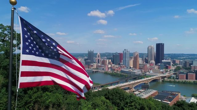 A slow push-in aerial view of an American flag on a flag pole above the Pittsburgh skyline on a summer day. As seen from Emerald View Park. Shot at 48fps for slow motion.  	