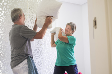 Fototapeta premium mature couple at home playing at the war with the pillows on the bed. morning activity for man and woman having fun. Happyness and foreverness life concept. white bedroom and laugh a lot