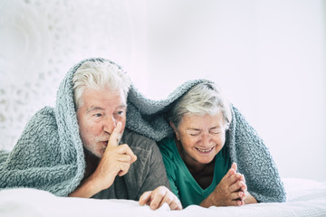 nice happy aged mature couple under the sheets whispering and asking for silence. the woman laugh a lot and both live happyness forever. togetherness concept for beautiful couple