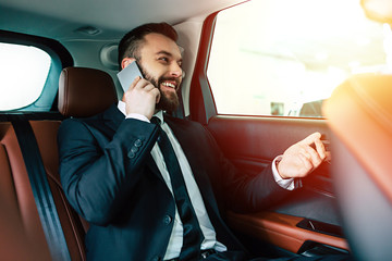 I'm on the road. Smiling businessman in full suit talking on phone while sitting on backseat of taxi car 