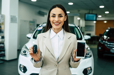 Beautiful smiling saleswoman in full suit in dealership on cars background with car keys and smart...
