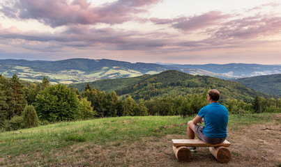 Fototapeta na wymiar A man is looking at a landscape of mountains, hills, meadow, forest during a cloudy sunset. Calm and peaceful looking at the beauty of a country.