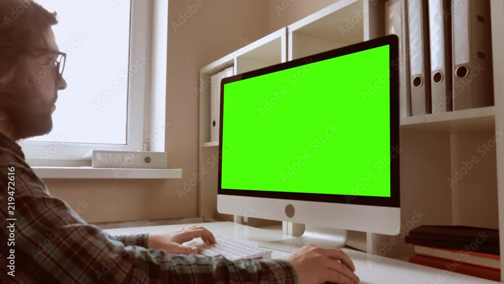 Poster man sitting at workplace paying game on computer in small office. businessman with beard looking on display with green screen. employee in startup company typing on keyboard use pc mouse
