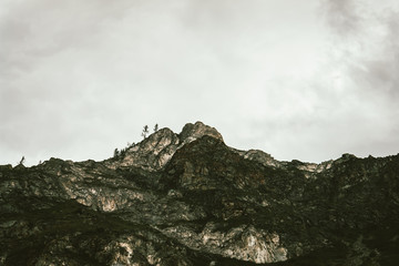 Rocky top of mountain with trees under cloudy sky close up. Steep slope. Gloomy overcast weather above wild. Minimalistic atmospheric tranquil mountain landscape of majestic nature.