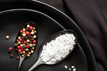 Close-up on salt and mixed pepper in dark spoons on black ceramic plate