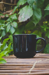 A lonf view of a blank black coffee mug on the wooden bamboo table in the park cafe under the over grown vines 