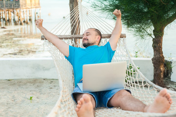 The young man lies in a hammock and is happy with a successful transaction. The concept of remote work