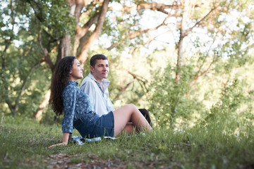 Naklejka na ściany i meble young couple sitting on the grass in the forest and looking on sunset, summer nature, bright sunlight, shadows and green leaves, romantic feelings
