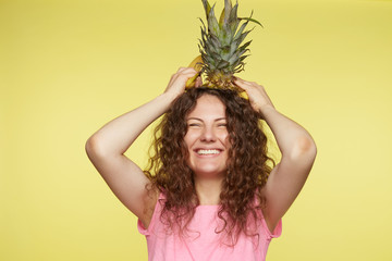 Joyful female with tanned skin, laughs happily, opens mouth widely, eyes closed, holds pineapple on head, has curly hair, isolated over yellow background. People, happiness and healthy lifestyle.