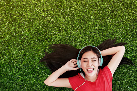 Happy Young Woman Lay on Green Grass to Listening Music via Headphone in the Park. Smiling and Closed Eyes, Top View