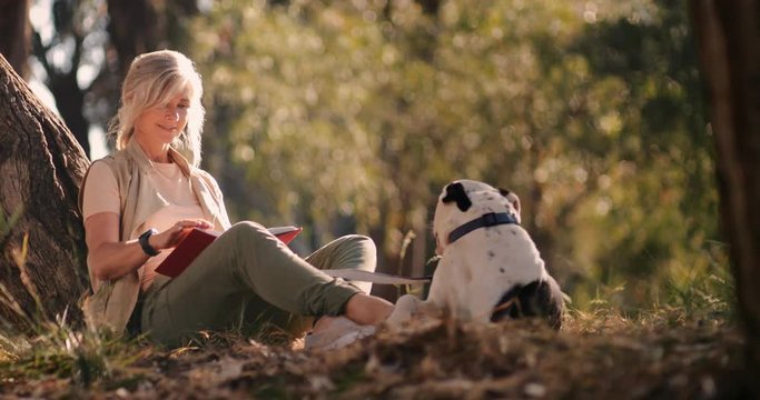 Happy senior woman with pet dog reading book in park
