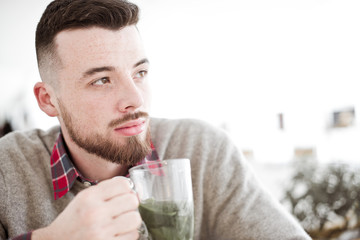 Fototapeta na wymiar Young handsome hipster man with cup of mint tea sitting in the room in cafe