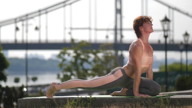 Mature redhead woman in sportswear practicing three-legged downward facing dog yoga pose in the morning in city. Fit, healthy beautiful middle-aged female working out near urban bridge.