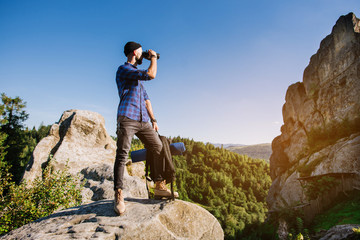 A traveler man standing on the rocks and holding backpack and binoculars over the blue sky background