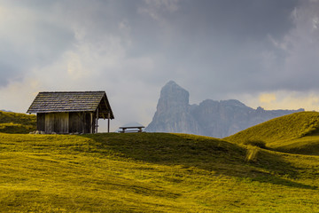 Old wooden shed with a bench on a top of the green grass hill with the mountain and cloudy sky in...