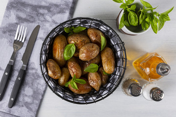 Baked potatoes in a black basket. Knife, fork, olive oil, salt, pepper, basil. The background is white. Top view. Action in the frame.