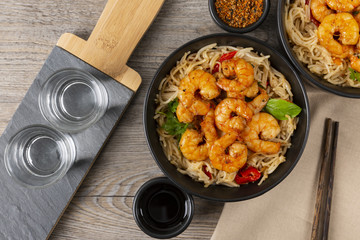 Fried prawns and rice noodles with spices on a black plate. Two glasses of vodka. Wooden background. Eastern food. Copy space. Horizontal shot.